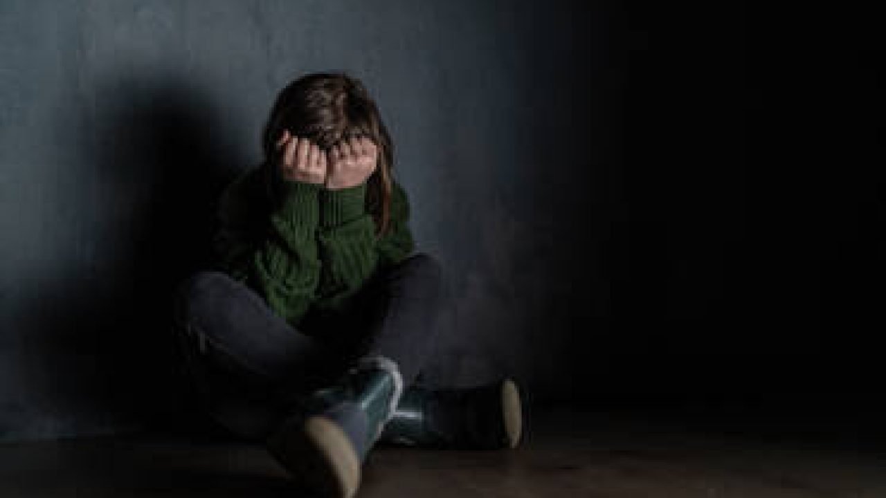 A sad little girl, alone in darkness, sitting and and covering face.